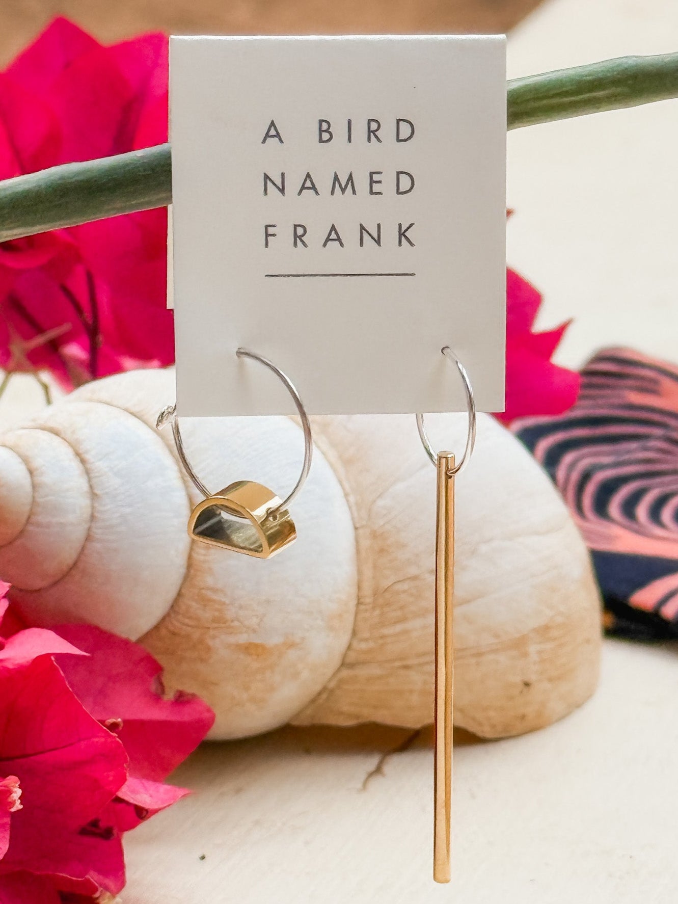 Brass and silver earrings on a card with 'A Bird Named Frank' text, surrounded by pink flowers and a shell.